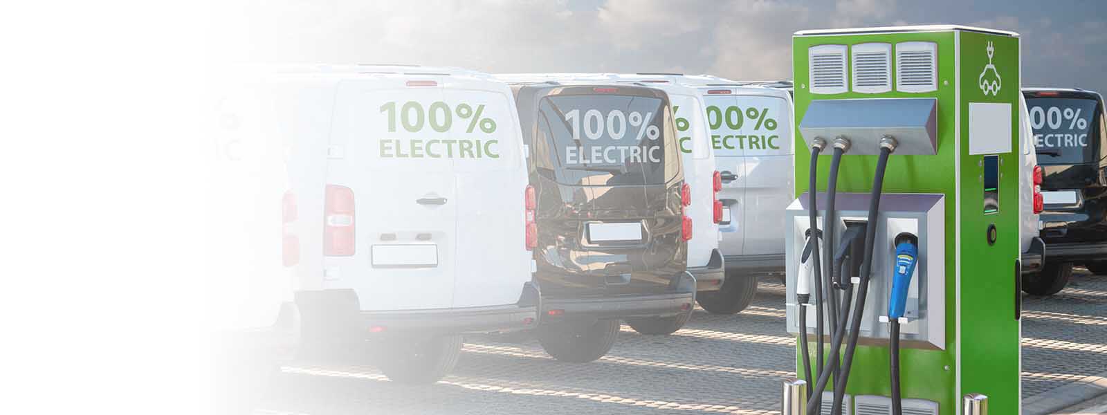 A fleet of vans that say “100% electric” are parked near an electric vehicle charging station