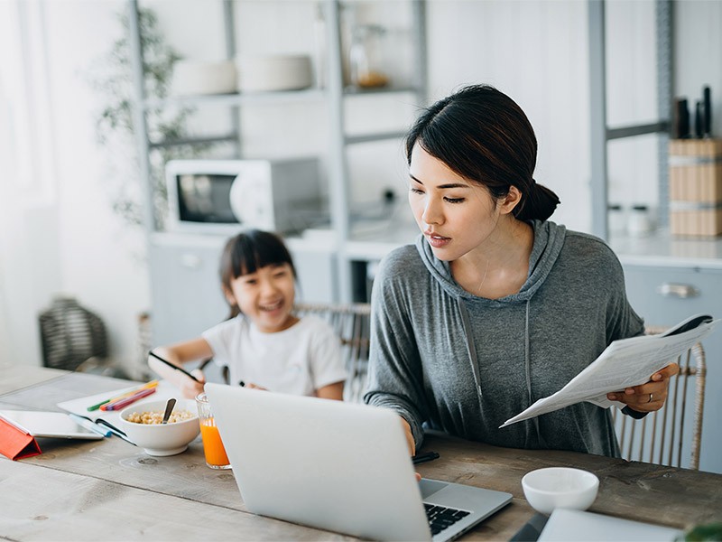 Young Asian mother working from home on a laptop while little daughter is studying from home